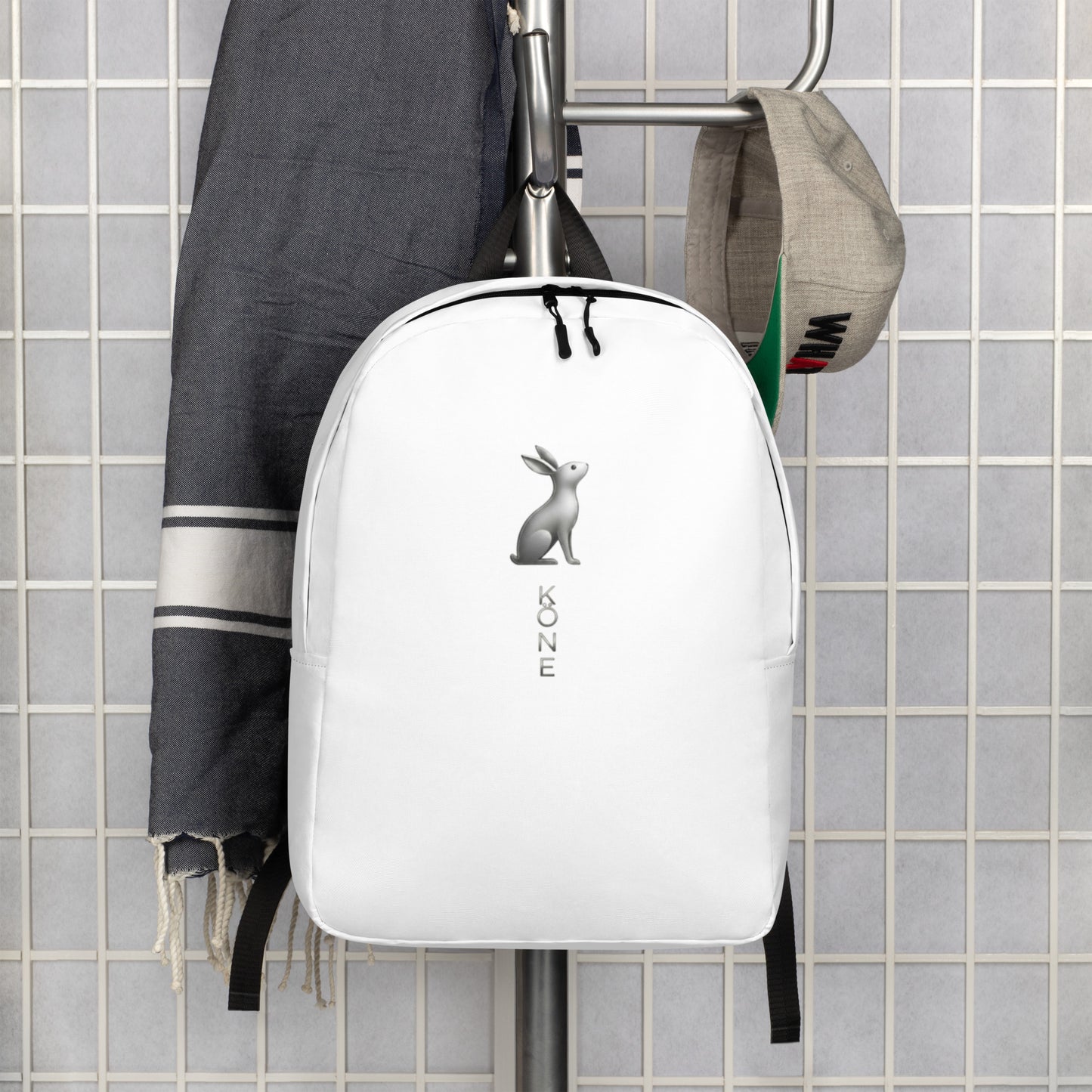 Mochila KÖNE blanca colgada junto a prendas urbanas, diseño minimalista y logo gris metalizado.