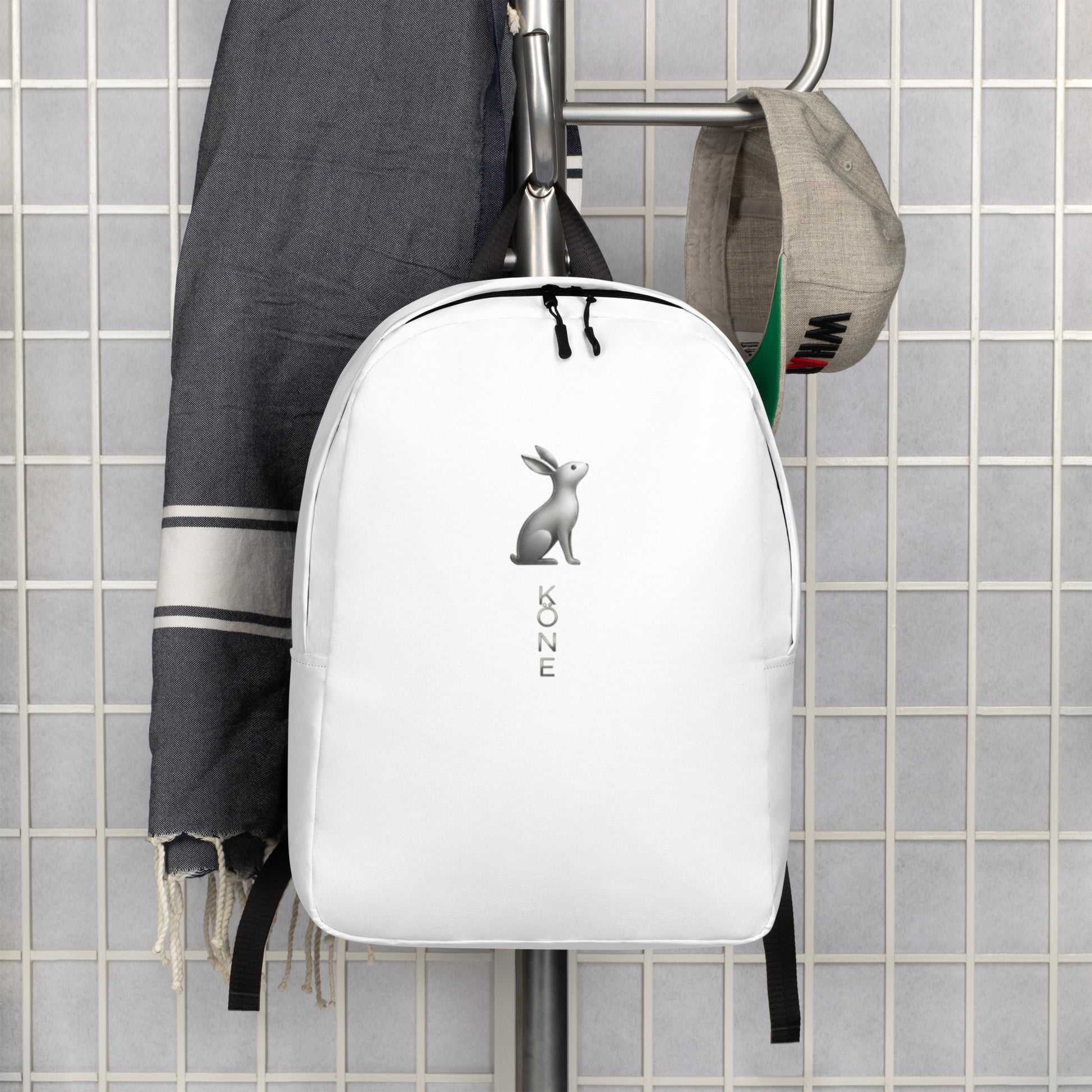Mochila KÖNE blanca colgada junto a prendas urbanas, diseño minimalista y logo gris metalizado.