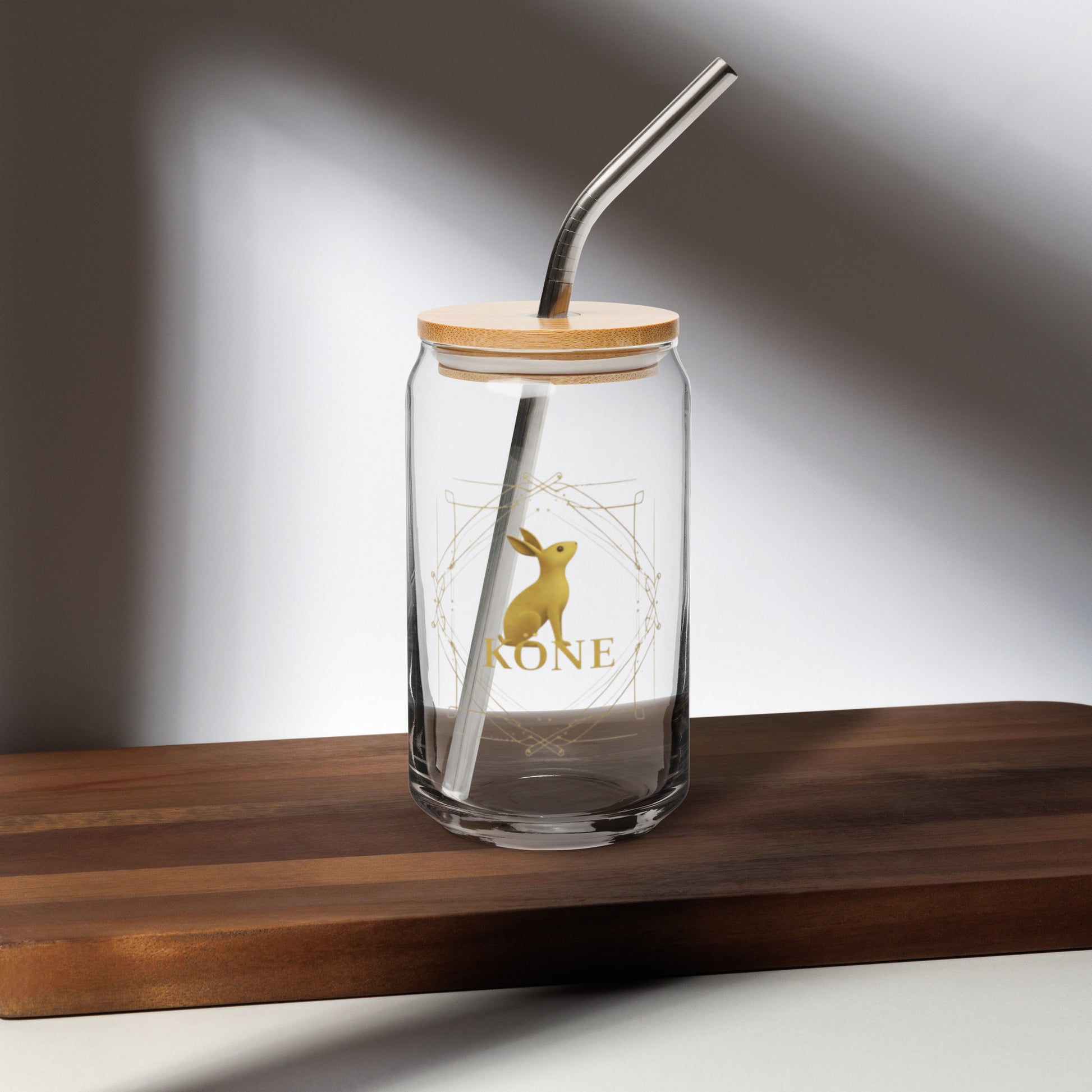 Vaso KÖNE con logo dorado, tapa de bambú y pajita metálica sobre mesa de madera con luz natural