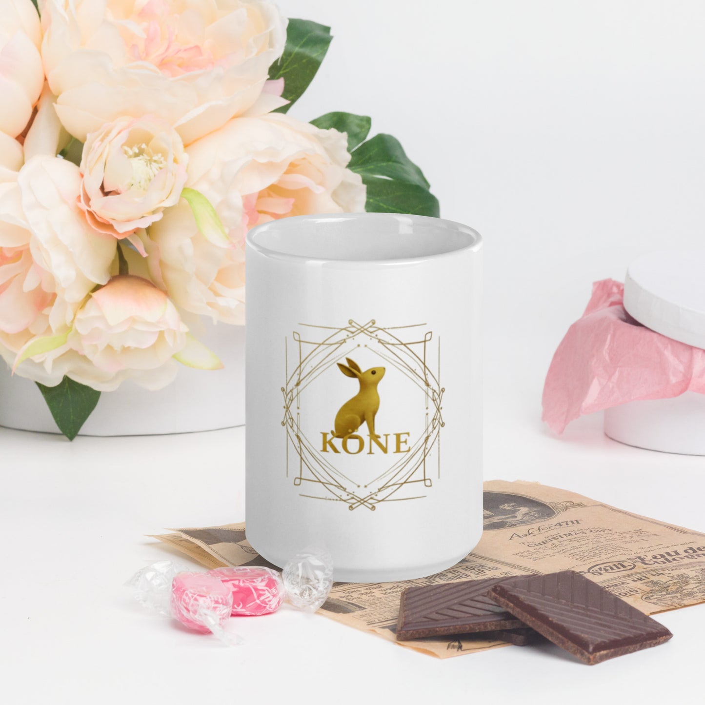 Taza KÖNE blanca con logo dorado ambientada en mesa de desayuno con flores y dulces.
