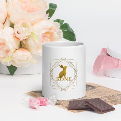 Detalle del logo dorado del conejo KÖNE en taza blanca sobre fondo floral claro.