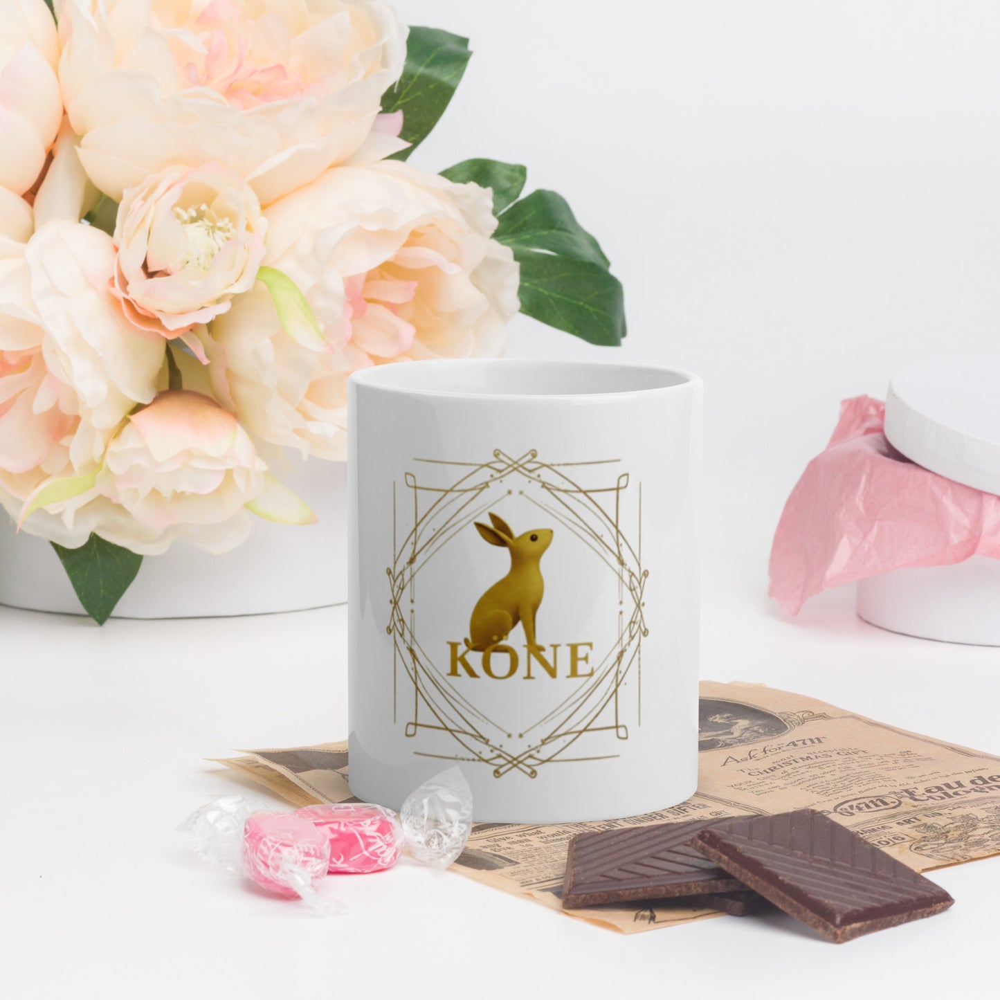 Taza KÖNE blanca con logo dorado en centro, colocada junto a flores rosadas y tableta de chocolate.