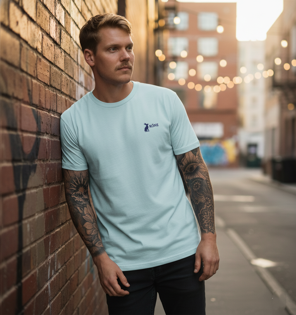 Camiseta KÖNE azul claro con logo bordado, modelo urbano apoyado en pared