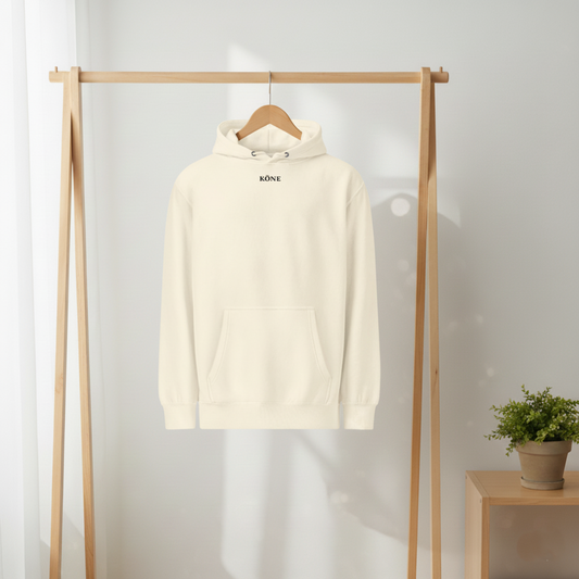 Sudadera KÖNE color crema colgada sobre perchero de madera, fondo beige minimalista.